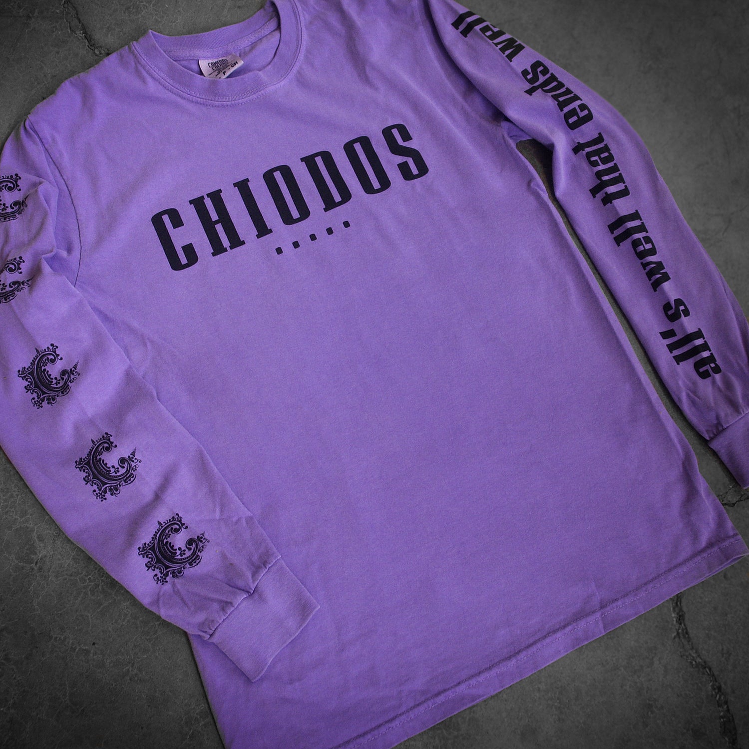 image of a violet long sleeve tee shirt laid flat on a concrete floor. small center chest print in black that says chiodos. left sleeve has the C logo. the right sleeve has a black print that says All's Well That Ends Well 