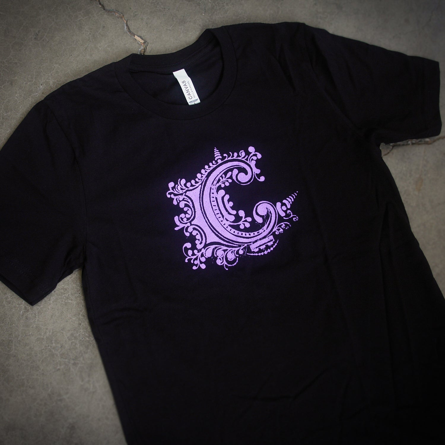 image of the front of a black tee shirt laid flat on a concrete floor. center chest print in purple of chiodos' C logo