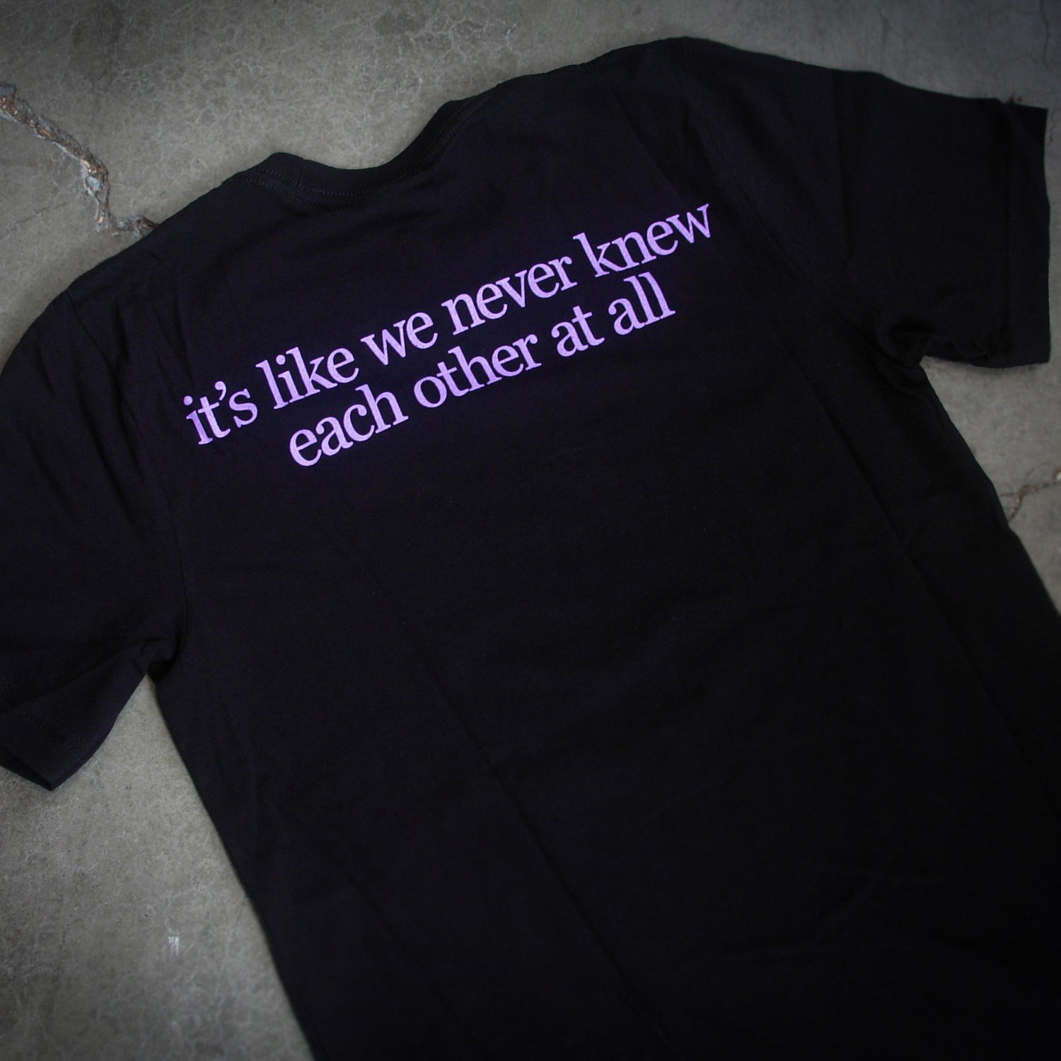 image of the back of a black tee shirt laid flat on a concrete floor. top center print in purple says it's like we never knew each other at all